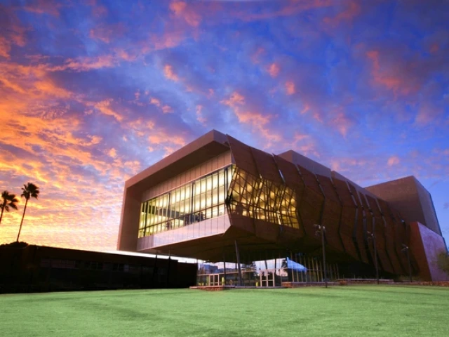 A modern, angular building with a geometric design situated against a dramatic sky during sunset. The structure features large glass windows framed by brown, asymmetric panels. The upper section of the building extends outward, creating an overhang supported by columns. The sky behind the building is a vibrant mix of orange and purple hues, with scattered clouds reflecting the sunset colors.
