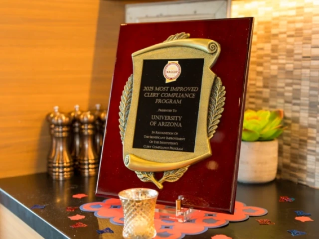 A close-up of a plaque reading "2025 Most Improved Clery Compliance Program, presented to University of Arizona," displayed on a countertop with small decorative items around it.