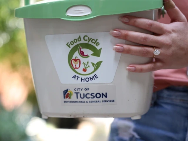A close-up of a person holding a small white composting bin labeled “Food Cycle at Home,” with logos for the City of Tucson Environmental & General Services. The person’s hand, wearing a large oval-cut engagement ring, rests on the bin.