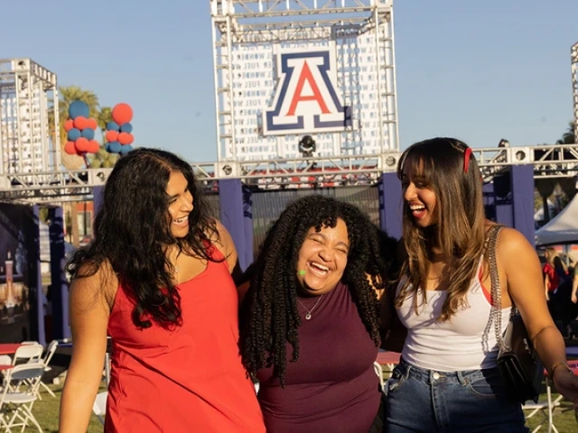 Three women smile and laugh in front of the Unviersity of Arizona logo.