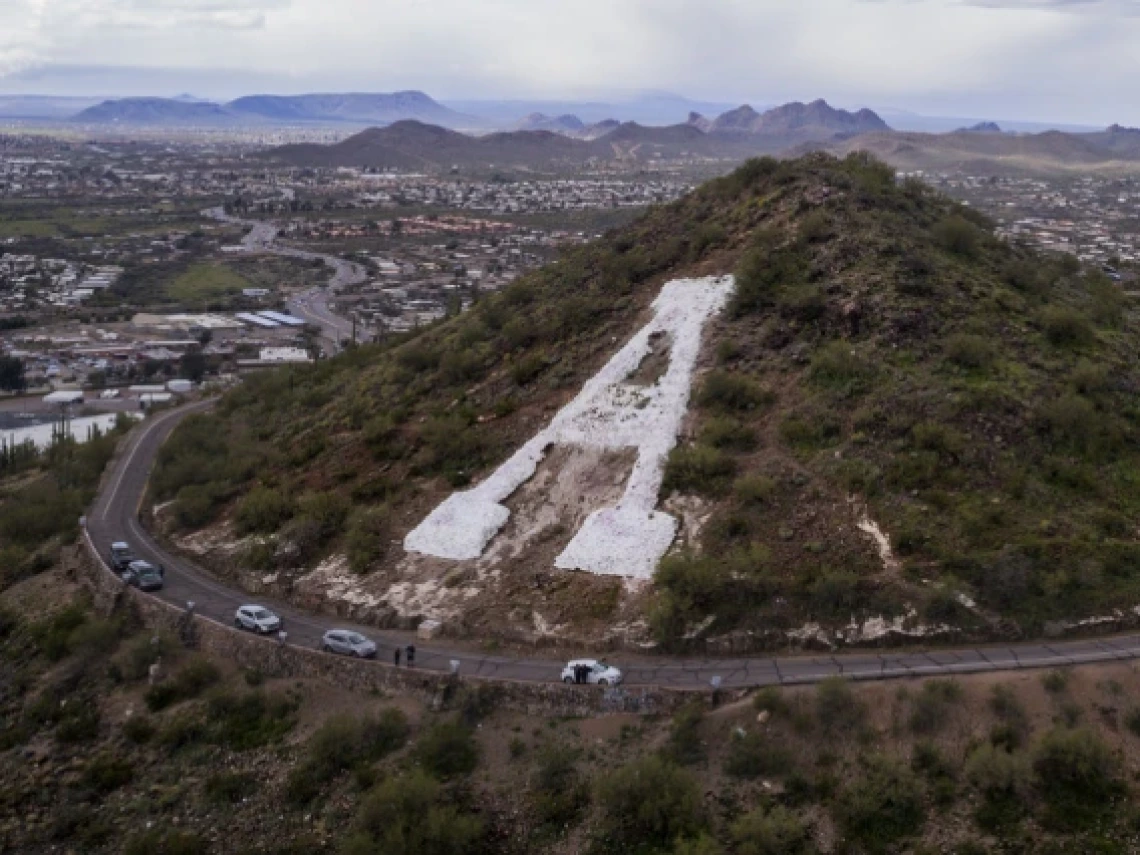 This image depicts a hillside with a large white letter "A" painted on it, surrounded by a winding road with several cars parked along the way. In the background, there is a vast cityscape extending into rolling desert mountains, under a partly cloudy sky.