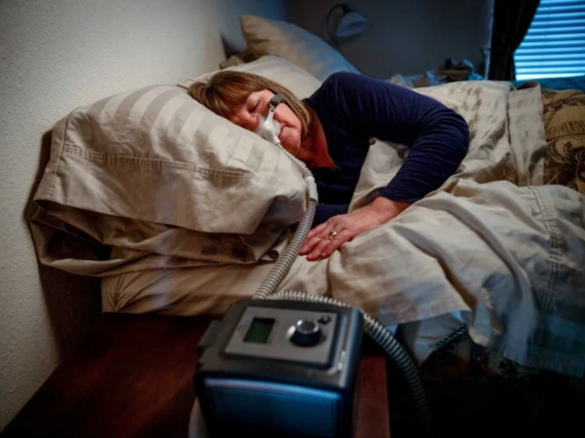 This image shows a woman sleeping in bed while using a CPAP (Continuous Positive Airway Pressure) machine for sleep apnea treatment. She is lying on her side with a nasal mask connected to the machine on a bedside table, surrounded by soft, patterned bedding.