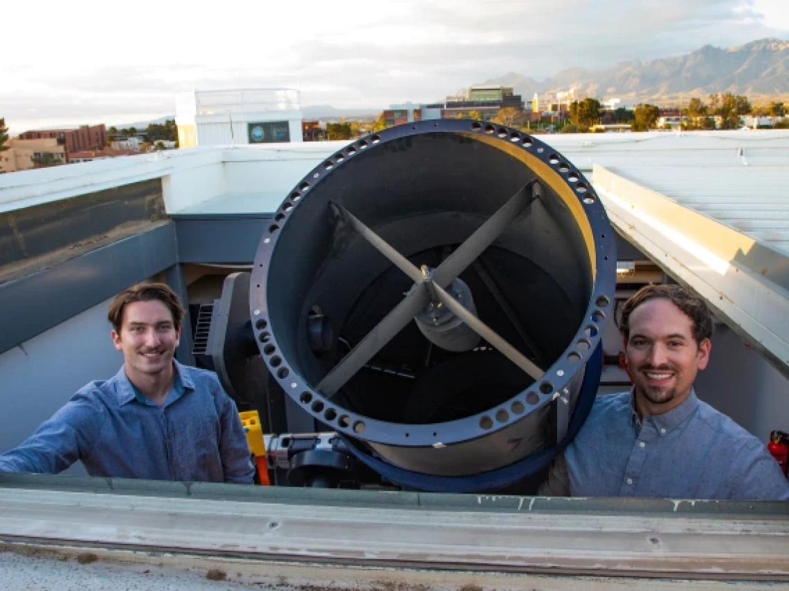 Two smiling men stand beside a large telescope on a rooftop observatory with a mountain range in the background. The image highlights the telescope's size and design, with the open sky and cityscape visible behind them, suggesting a setting for astronomical research.