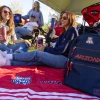 People relax under a canopy at a tailgate on a sunny day, sitting on a red University of Arizona blanket. A navy backpack with “Arizona” and the “A” logo is in the foreground.